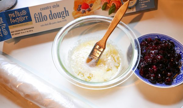 A table top with a Fillo Factory Box of fillo dough, a bowl of butter and some blueberry filling in a bow.